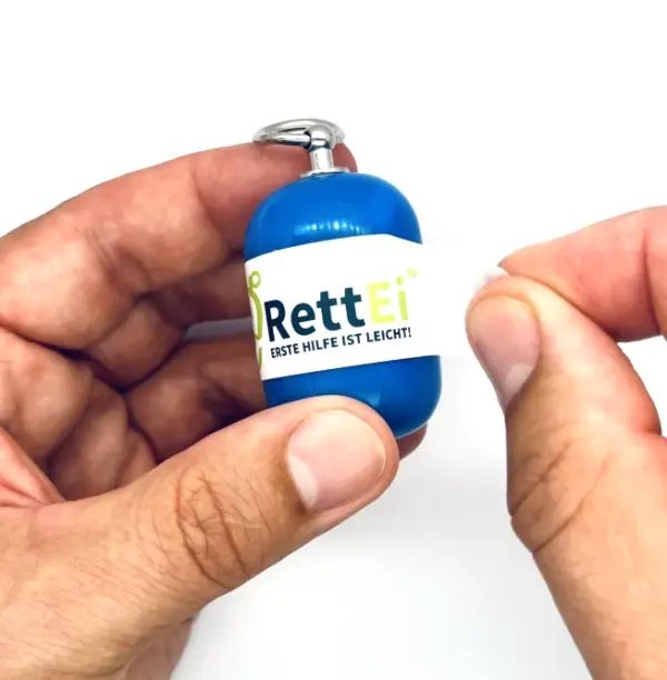 Notfall-Set-fuer-unterwegs-als-RettEi-Original-via-Erste-Hilfe-Schluesselanhaenger-mit-Beatmungstuch-zur-Mund-zu-Mund-Beatmung-mit-blauer-Kapsel-beim-oeffnen.
