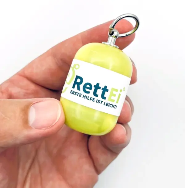 Notfall-Set-fuer-unterwegs-als-RettEi-Original-via-Erste-Hilfe-Schluesselanhaenger-mit-Beatmungstuch-zur-Mund-zu-Mund-Beatmung-mit-gelb-Kapsel-in-Hand.