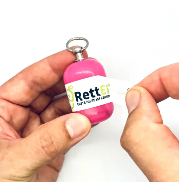 Notfall-Set-fuer-unterwegs-als-RettEi-Original-via-Erste-Hilfe-Schluesselanhaenger-mit-Beatmungstuch-zur-Mund-zu-Mund-Beatmung-mit-rosa-Kapsel-beim-oeffnen.