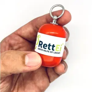 Notfall-Set-fuer-unterwegs-als-RettEi-Original-via-Erste-Hilfe-Schluesselanhaenger-mit-Beatmungstuch-zur-Mund-zu-Mund-Beatmung-mit-roter-Kapsel-in-Hand