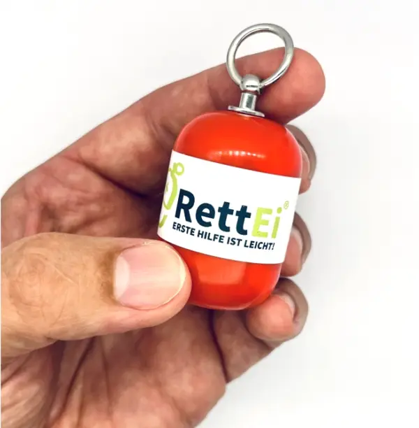 Notfall-Set-fuer-unterwegs-als-RettEi-Original-via-Erste-Hilfe-Schluesselanhaenger-mit-Beatmungstuch-zur-Mund-zu-Mund-Beatmung-mit-roter-Kapsel-in-Hand