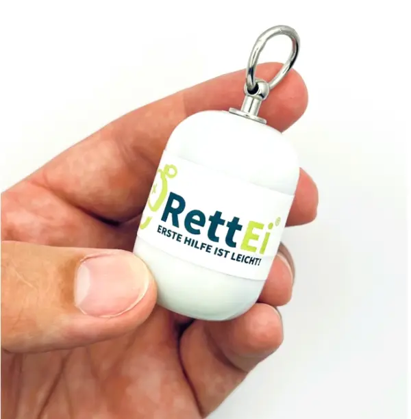 Notfall-Set-fuer-unterwegs-als-RettEi-Original-via-Erste-Hilfe-Schluesselanhaenger-mit-Beatmungstuch-zur-Mund-zu-Mund-Beatmung-mit-weiss-Kapsel-in-Hand.
